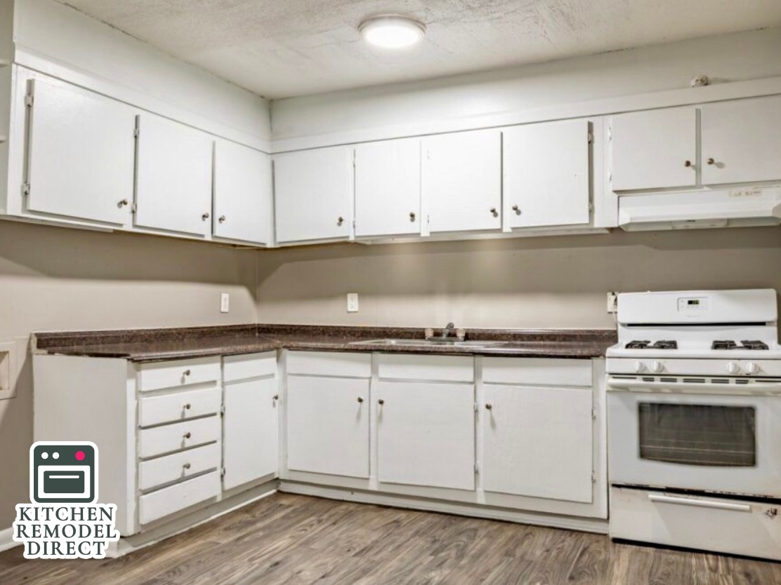 A bright kitchen featuring white painted cabinets with small dark knobs and a dark, speckled brown countertop. The space includes a white electric stove and range hood, dark wood-laminate flooring, and a single flush-mount ceiling light. A "Kitchen Remodel Direct" logo is in the bottom left corner.