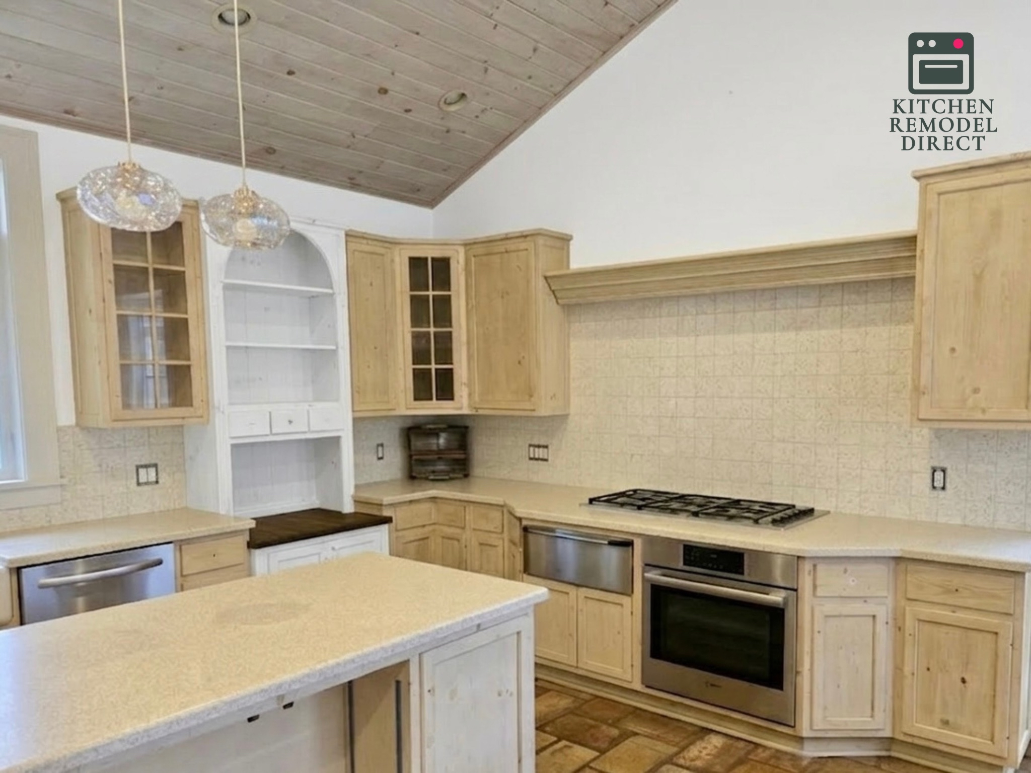 A modern rustic kitchen featuring light wood cabinetry and a vaulted ceiling with wood planks. The space includes a stainless steel range and oven, a neutral tiled backsplash, and light-colored stone countertops. Two glass pendant lights hang over a central island, and a white arched hutch is built into the wall. A "Kitchen Remodel Direct" logo is in the top right corner.
