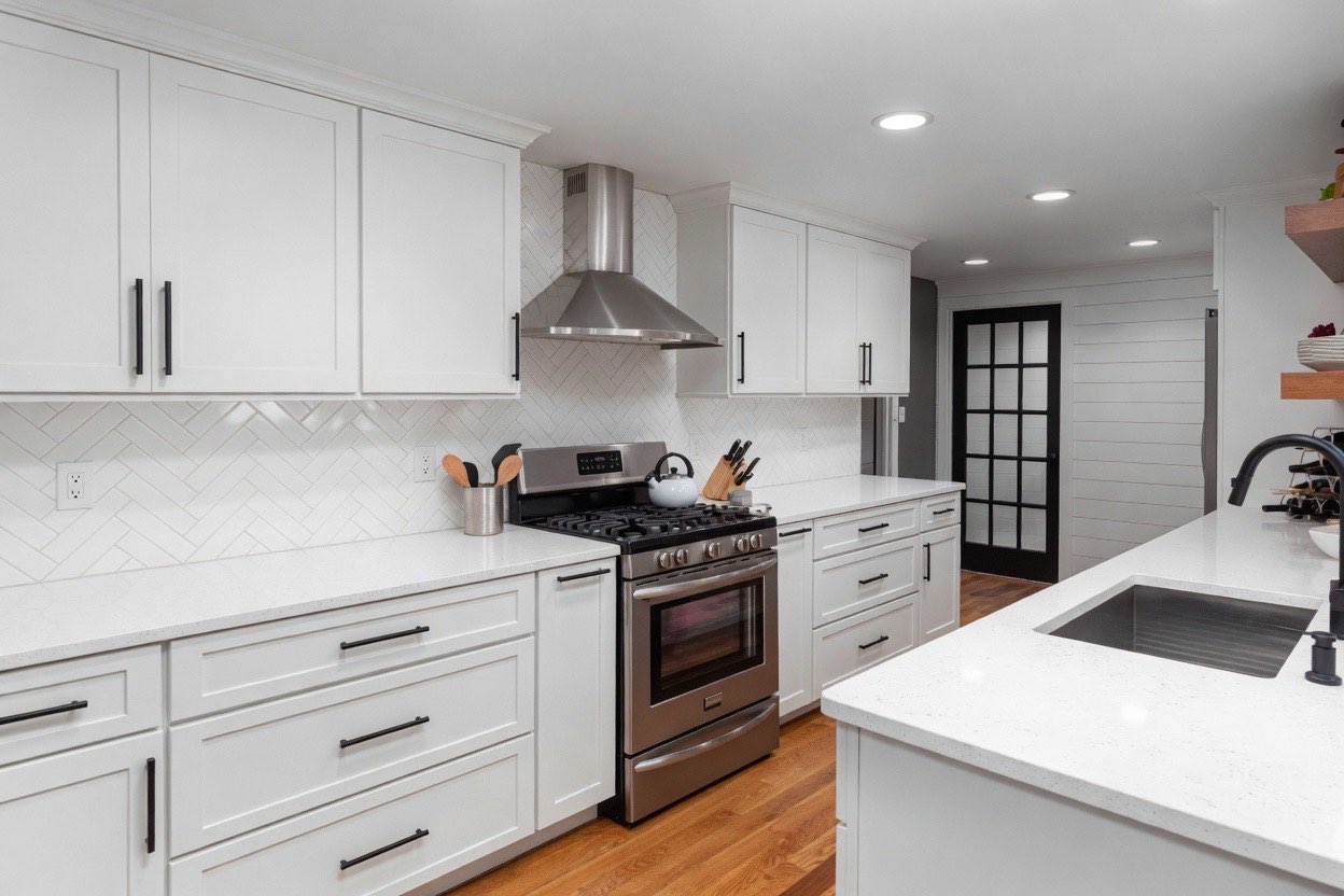 A high-contrast, modern kitchen corner featuring white shaker-style cabinets with brushed nickel handles and light marble-patterned countertops. The space includes a stainless steel French door refrigerator, an LG glass cooktop, and a marble-look tiled floor. A small coffee station with an espresso machine and coffee canisters sits against a gray and white mosaic tile backsplash.