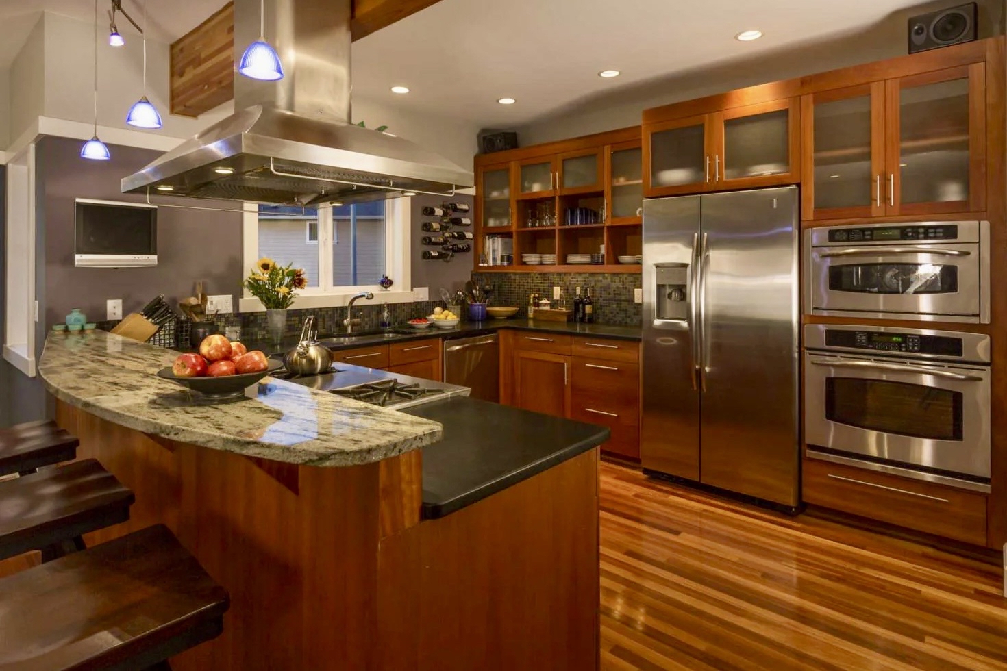 Modern kitchen featuring warm medium-toned wood cabinetry, frosted glass upper cabinet doors, and polished hardwood floors. A large stainless steel range hood hangs over a peninsula with a curved upper granite breakfast bar and dark barstools. High-end stainless steel appliances include a side-by-side refrigerator and double wall ovens, set against a dark tiled backsplash and gray accent wall.