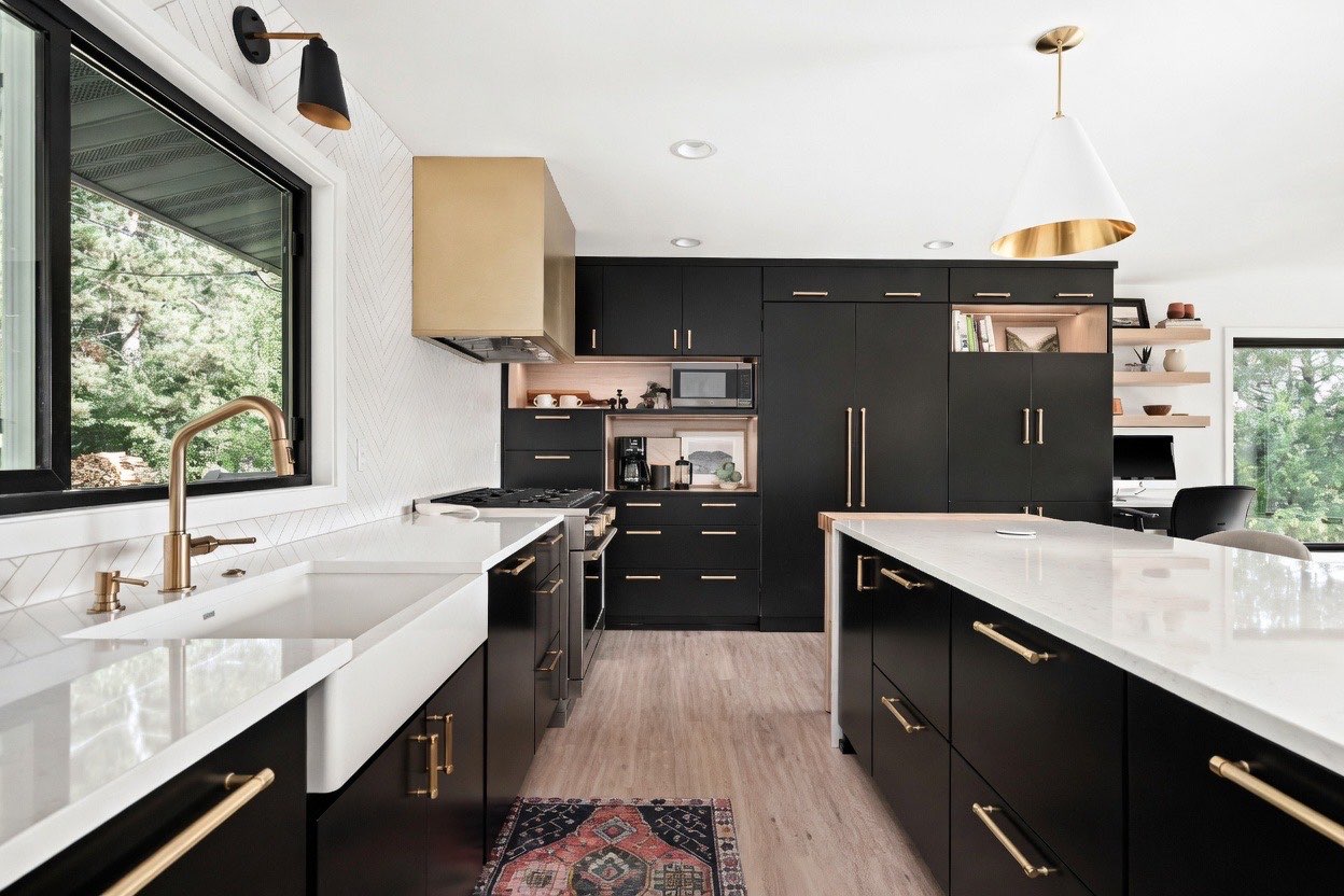 Modern, high-contrast kitchen with matte black cabinetry, gold hardware, and white quartz countertops. A large white apron-front sink with a brushed gold faucet sits beneath a large black-framed window on the left, next to a wall with white chevron tile backsplash. A metallic gold range hood hangs over a gas stove, while a large black kitchen island with a white cone pendant light overhead stands in the foreground on light wood floors.